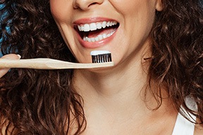 Nose-to-shoulder view of woman smiling holding wooden toothbrush with toothpaste to her teeth